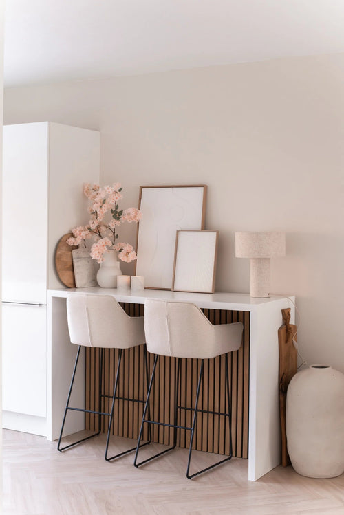 A minimalist home bar with two beige stools with cushions, a white counter, framed art, a table lamp, neutral toned decoration and Artificial Cherry Blossom 100 cm light pink in vases against a light-colored wall.