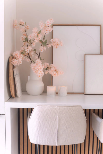 A minimal desk setup with a white chair, two candles, decorative trays, framed abstract art and an Artificial Cherry Blossom 100cm light pink in a white vase on a white surface.