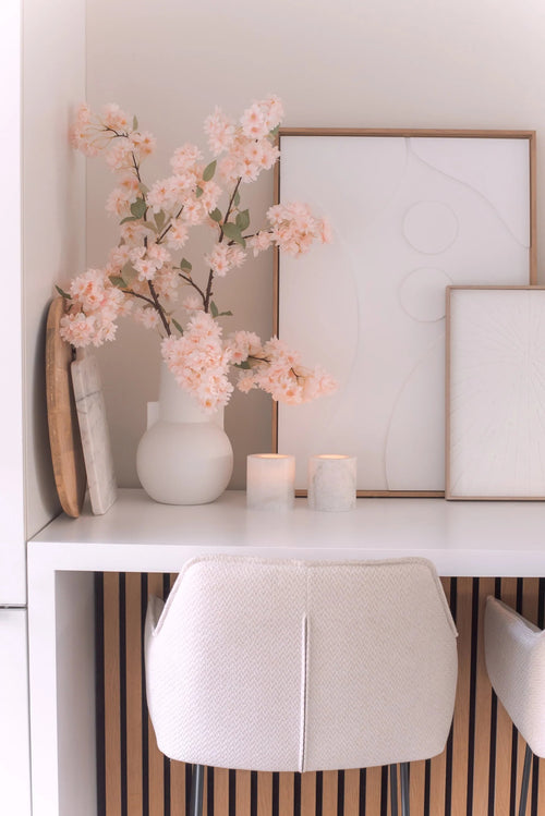 A minimal desk setup with a white chair, two candles, decorative trays, framed abstract art and an Artificial Cherry Blossom 100cm light pink in a white vase on a white surface.
