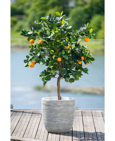 A 120 cm artificial orange tree with ripe fruits stands in a white embossed pot on a wooden terrace, overlooking a hazy water feature and a green landscape outside.