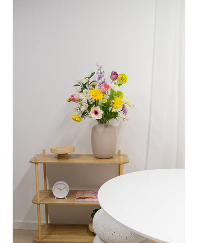 A light wooden shelf shows a beige vase with Artificial Ranunculus Real Touch Yellow 60cm flowers, a white clock and a wooden tray. Part of a round white table appears in the foreground against a plain white wall.