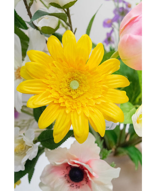The Artificial Flower Gerbera 53 cm yellow, a vibrant yellow artificial flower with layered petals, stands among other colorful artificial flowers and green leaves, with white and pink flowers in the background.