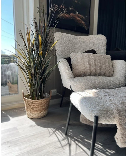 A cozy corner with a white fluffy armchair, matching ottoman, textured cushion and the Artificial Grass Bordeaux UV 120cm in a woven basket with sunlight shining through a large window.