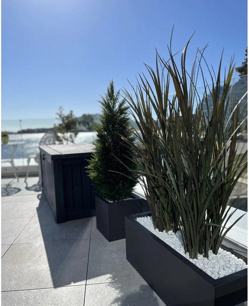 Artificial grass Bordeaux UV 120cm in rectangular planters with white stones decorates a modern outdoor terrace against a background of blue sky and trees in the distance.