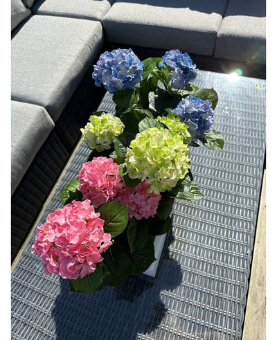 The Artificial Hydrangea Green UV 35 cm plant stands on a wicker table with a glass top next to a gray outdoor sofa, with realistic green hydrangeas that light up in the sunlight.