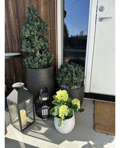 An Artificial Hydrangea Green UV 35 cm plant in a white pot with yellow-green flowers stands on the veranda near a brown door, next to two plants in a black pot and two decorative lanterns with candles.