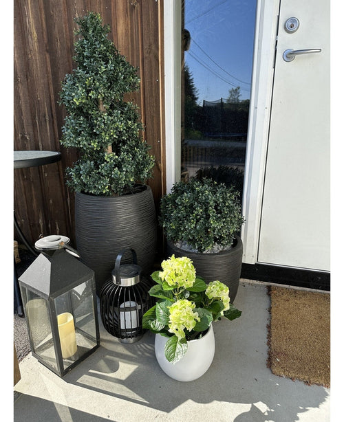 An Artificial Hydrangea Green UV 35 cm plant in a white pot with yellow-green flowers stands on the veranda near a brown door, next to two plants in a black pot and two decorative lanterns with candles.