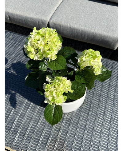 The Artificial Hydrangea Green UV 35 cm, with light green blossoms and dark green leaves, stands on a woven gray outdoor table with a cushioned bench in the background.