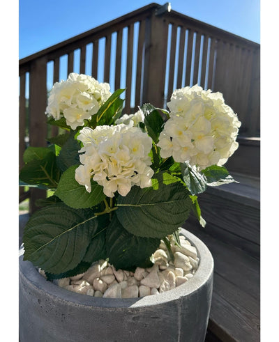 The Artificial Hydrangea White UV 35 cm plant, with white flowers and green leaves, stands outside on white pebbles on a sunny day, with wooden patio railings and a staircase in the background.