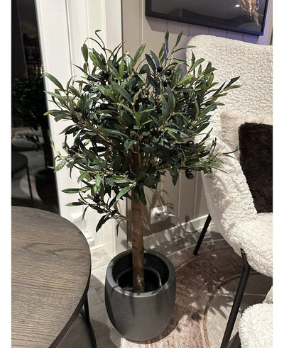 The Artificial olive tree UV 85 cm, a compact and easy-care artificial plant with dark green leaves and black olives, stands indoors next to a round wooden table and an armchair with a white texture.