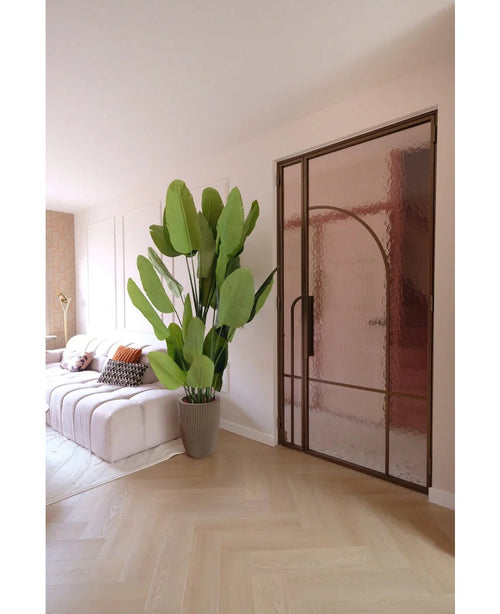 A modern living room with a white sofa, patterned cushions and the Artificial Plant Strelitzia Real Touch Deluxe 210 cm. A light wooden floor, soft pink walls and a glass door with a bronze metal frame create a light, airy atmosphere.