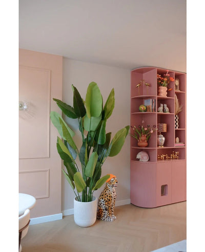 The Artificial Plant Strelitzia Real Touch Deluxe 210 cm stands next to a ceramic cheetah figure with an orange hat, next to a high pink shelf full of vases, flowers and ornaments in a pastel-colored room.