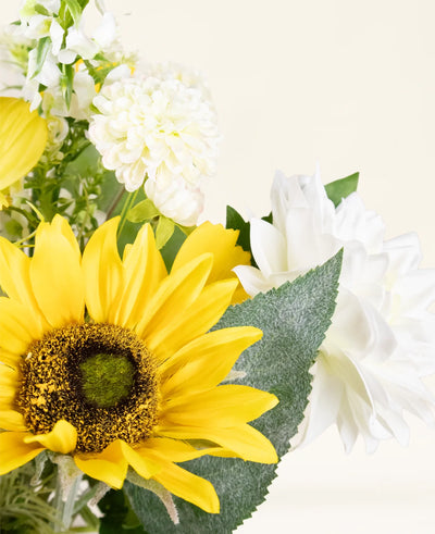 A close-up of the Sunny Whisper silk artificial flower bouquet with a bright yellow sunflower, white dahlias and green leaves against a light, neutral background.