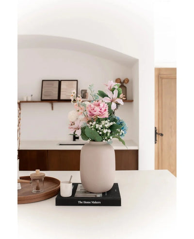 The Vase Artic Beige Ø21 x H29 cm, filled with pink and blue artificial flowers, stands on a black book on a white countertop. Soft lighting accentuates the minimalist scene with decor shelves and a wooden door in the background.