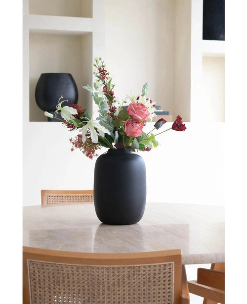 The Vase Artic Black Ø21 x H29 cm, filled with pink, white and red flowers, stands on a round beige table surrounded by two wooden chairs with woven wicker backrests. Decorative planks are visible in the background.