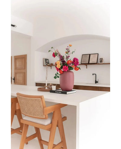 A modern kitchen with a white island, a wooden chair and the Artic Pink Vase Ø21 x H29 cm filled with colorful flowers. In the background you see shelves with books and decor, a tray and a black tap.