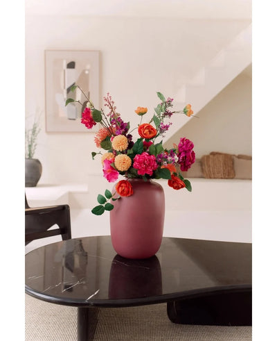 The Vase Artic Pink Ø21 x H29 cm, filled with pink, orange and red flowers, stands on a dark glossy coffee table in a modern minimalist living room with a staircase and framed works of art in the background.