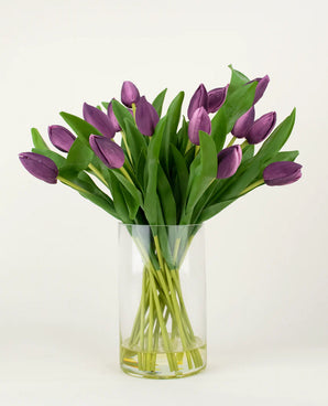 The Silk artificial bouquet Tulips in vase Purple has long-stemmed purple artificial tulips with lush green leaves, beautifully arranged in a clear glass vase against a plain white background.