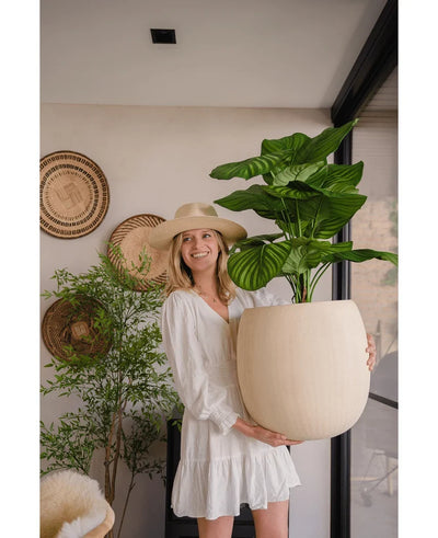 A woman in a white dress and hat smiles as she holds the Artificial Calathea UV 72 cm plant indoors, with woven baskets and another plant in the background.