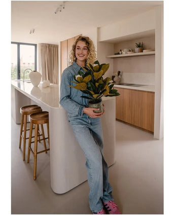 A smiling person in denim clothes stands in a modern kitchen holding an Artificial Magnolia plant in a 46 cm pot, an easy-care plant. The kitchen, which is perfect for interior decoration, features wooden cabinets, a white kitchen island and two bar stools.