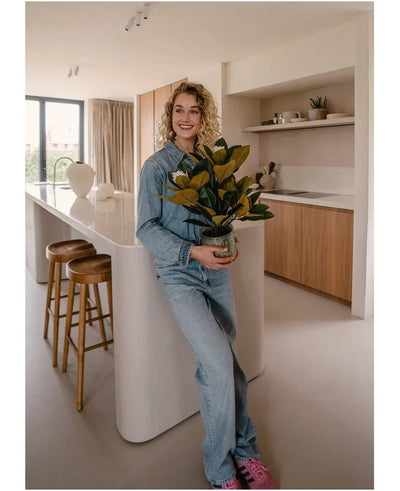 A smiling person in denim clothes stands in a modern kitchen holding an Artificial Magnolia plant in a 46 cm pot, an easy-care plant. The kitchen, which is perfect for interior decoration, features wooden cabinets, a white kitchen island and two bar stools.