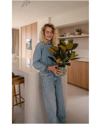 A woman with curly blond hair, dressed in denim clothes, stands smiling in a modern kitchen with the Artificial Magnolia plant in a 46 cm pot in her hands, surrounded by wooden cupboards and shelves – an ideal choice for anyone looking for low-maintenance plants to enhance their stylish interior.