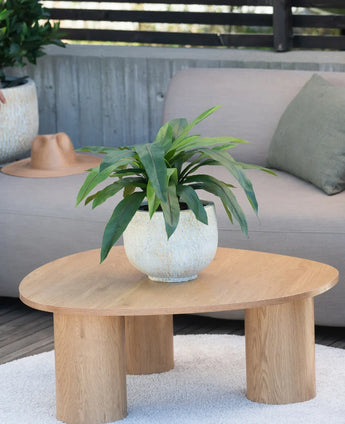 An artificial plant Bird's Deluxe of 60 cm stands on a round wooden coffee table in front of a beige sofa, decorated with a green cushion and a brown hat hanging over the armrest.