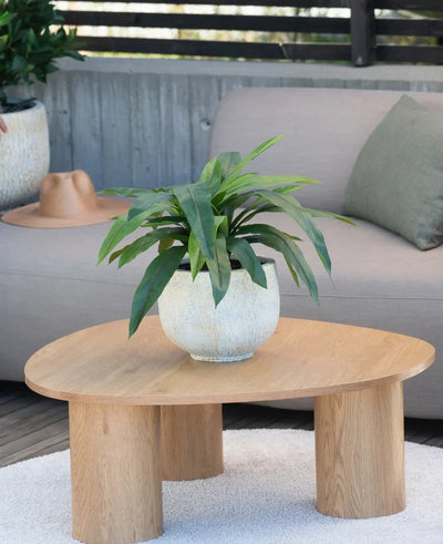 An artificial plant Bird's Deluxe of 60 cm stands on a round wooden coffee table in front of a beige sofa, decorated with a green cushion and a brown hat hanging over the armrest.