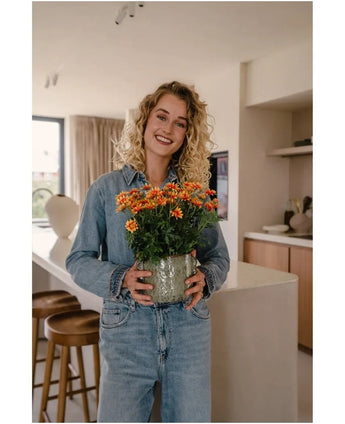 A woman with curly blond hair, wearing denim clothes, smiles as she holds the Artificial Daisy UV 31 cm plant – a pot of bright orange and yellow flowers – in a modern kitchen.