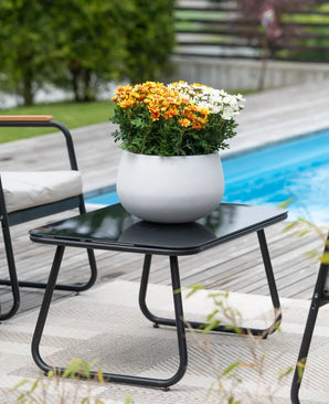 A black patio table with an Artificial Daisy UV 31 cm plant – a round white pot with vibrant orange and white daisies – stands next to a swimming pool, surrounded by garden chairs and with greenery in the background.