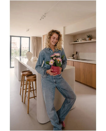 Een vrouw in spijkerbroek staat in een moderne keuken, glimlachend en met de Kunstplant Gerbera UV 28 cm roze in haar handen – een kleine roze bloempot met roze en witte bloemen. Op de achtergrond zijn twee houten krukken en een groot raam te zien.