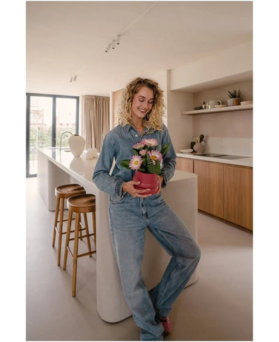Een vrouw in spijkerbroek staat in een moderne keuken, glimlachend en met de Kunstplant Gerbera UV 28 cm roze in haar handen – een kleine roze bloempot met roze en witte bloemen. Op de achtergrond zijn twee houten krukken en een groot raam te zien.
