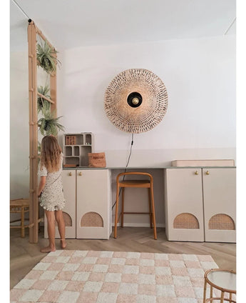 Een jong meisje in een lichte jurk staat naast een plank waarop de Kunst-hangplant ‘Frosted Varen op bal 76 cm’ en andere hangplanten staan, in een moderne kamer met beige kasten, een hoge kruk, een wandlamp van riet en een geruit vloerkleed op de vloer.