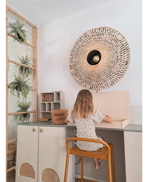 Een jong kind met schouderlang haar zit aan een bureau in een lichte kamer, met de rug naar de kijker gekeerd. Achter haar staan een geweven wandlamp, enkele kasten en de Kunst-hangplant ‘Frosted Varen op bal 76 cm’, genesteld tussen de planten op een houten ladder, naast kleine plankjes met netjes gerangschikte spullen.