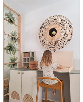 Een jong kind met schouderlang haar zit aan een bureau in een lichte kamer, met de rug naar de kijker gekeerd. Achter haar staan een geweven wandlamp, enkele kasten en de Kunst-hangplant ‘Frosted Varen op bal 76 cm’, genesteld tussen de planten op een houten ladder, naast kleine plankjes met netjes gerangschikte spullen.