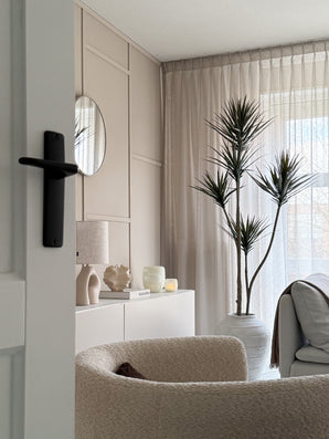 Een moderne, minimalistische woonkamer met een beige fauteuil met textuur, Kunstplant Dracaena Fragnans 180 cm, wit dressoir met decor, ronde spiegel en transparante gordijnen die zacht daglicht filteren door het raam.