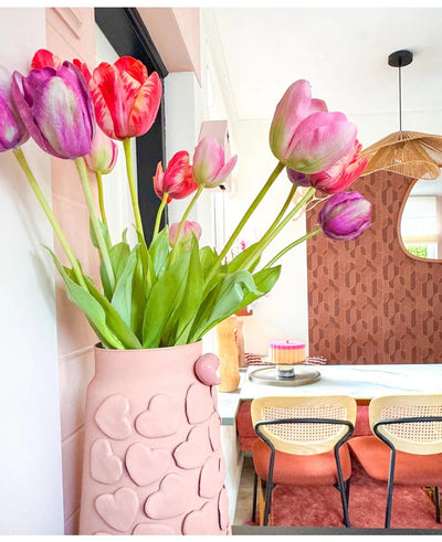 Een roze vaas met hartjes in reliëf houdt de Kunstbloem Franse Tulp 60 cm rood op een eettafel. Daarachter staan stoelen met rieten ruggen, een taart op tafel en een spiegel op een gedessineerde bruine muur onder een geweven hanglamp.