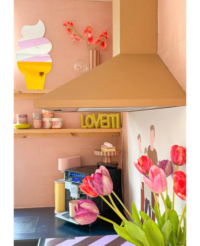 Een pastelroze keuken met Kunstbloem Franse Tulp 60 cm paars op de voorgrond, een geel LOVETTI bord op de plank, roze mokken, muurkunst van een ijshoorntje en een minimalistische muurschildering van twee mensen op de muur.