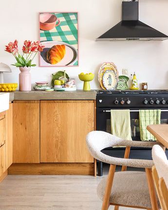 Een lichte keuken met houten kasten, een zwart fornuis, groene handdoeken, kleurrijk servies en een ingelijste afbeelding van een gebakje op een geruite tafel krijgt een frisse uitstraling dankzij de Kunstbloem Franse Parkiet Tulp 76 cm rood/geel. Een gezellige witte stoel staat naast een licht houten eettafel.