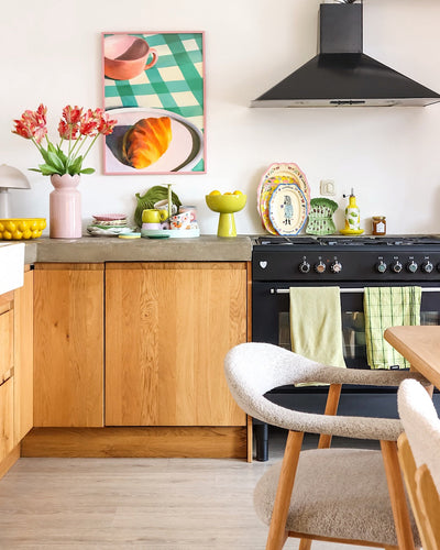 Een lichte keuken met houten kasten, een zwart fornuis, groene handdoeken, kleurrijk servies en een ingelijste afbeelding van een gebakje op een geruite tafel krijgt een frisse uitstraling dankzij de Kunstbloem Franse Parkiet Tulp 76 cm rood/geel. Een gezellige witte stoel staat naast een licht houten eettafel.
