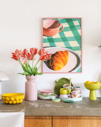 Een aanrecht met allerlei kleurrijke serviesstukken en een Kunstbloem Franse Parkiet Tulp van 76 cm in rood/geel in een roze vaas. Daarboven hangt een ingelijste afbeelding van een croissant en een theekopje op een groen geruit tafelkleed.