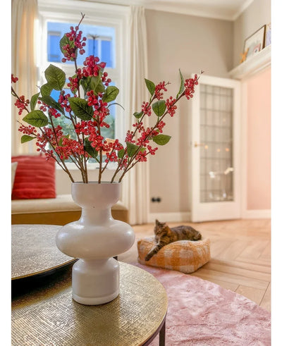 On a table is a white vase with pink Kunsttak berries (64 cm) and green leaves. In the background, a cat lies on a pink cushion in a bright living room with beige walls and a glass door.