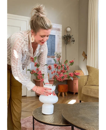 A woman with blond hair in a bun arranges the Kunsttak Berries 64 cm pink in a white vase on a round table in a cozy living room with earth tones and modern furnishings.
