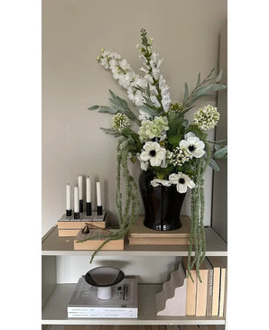 On a shelf against a light wall there is a black vase with the Artificial Flower Amaranthus 124 cm green, next to five white candles, beige books and decorative objects in neutral tones.