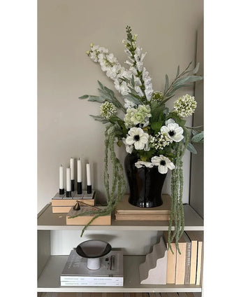 On a shelf against a light wall there is a black vase with the Artificial Flower Amaranthus 124 cm green, next to five white candles, beige books and decorative objects in neutral tones.