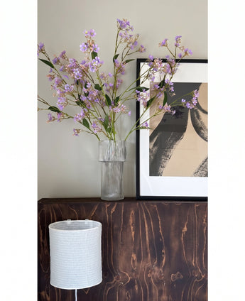 In een doorzichtige vaas staan de bloemen van de Kunsttak Daphne Spray 108 cm paars op een donkerhouten oppervlak naast een modern, abstract kunstwerk in een lijst, met daaronder een witte lampenkap met structuur.