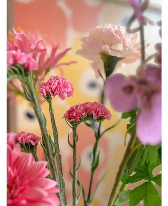 Een close-up van de Kunsttak Statice 64 cm roze kunstbloemen, met delicate roze statice-bloemen tegen een wazige, pastelkleurige achtergrond met abstracte gele en perzikkleurige vormen.