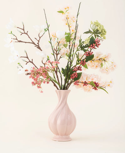 Een roze vaas van keramiek bevat het Zijden kunstbloemen boeket Springtime Poem, met witte, roze en groene bloemen met groene bladeren en bruine stelen, tegen een lichte achtergrond.