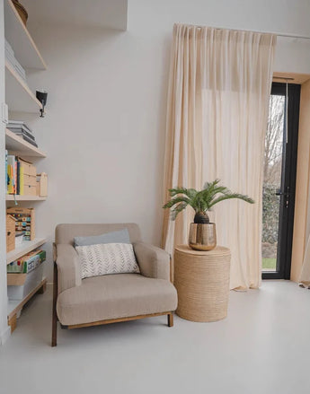 A cozy reading corner with a beige armchair, a round wicker side table with the Artificial Baby Cycas Palm 60 cm plant on it, bookcases and net curtains that hang over a glass door that opens onto the garden.