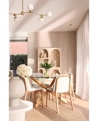 A modern dining area has a round glass table, wooden chairs with white cushions and the Silk artificial bouquet Tulips in vase White as the centrepiece. Decorative shelves with vases and transparent curtains complete the beige space.